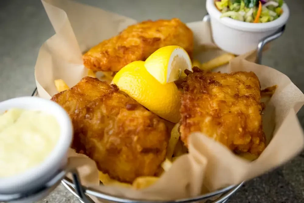 Crispy fish and chips, lemon, tartar, and salad in a basket.