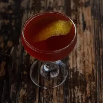 Dark red-brown cocktail in coupe with twisted citrus peel on wood.