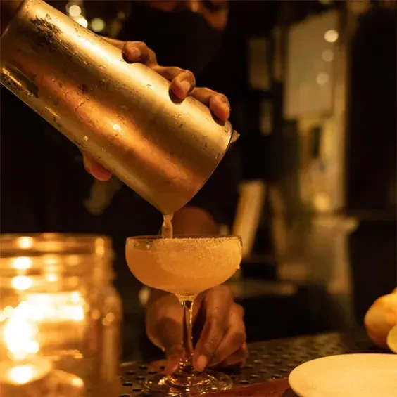 Hands pour pale yellow drink from shaker into sugared glass at bar.