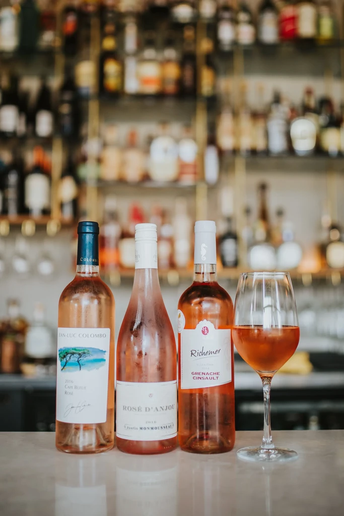 Three rosé wine bottles and glass at a well-stocked bar.