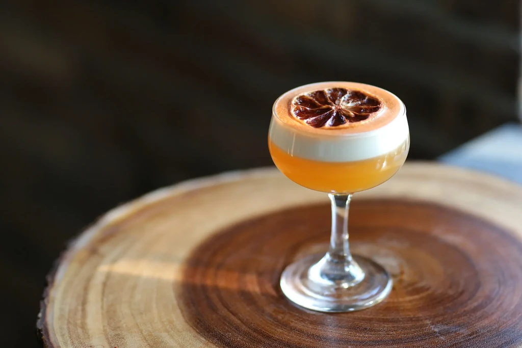 Three-layered cocktail with orange, white foam, dried citrus on wood.