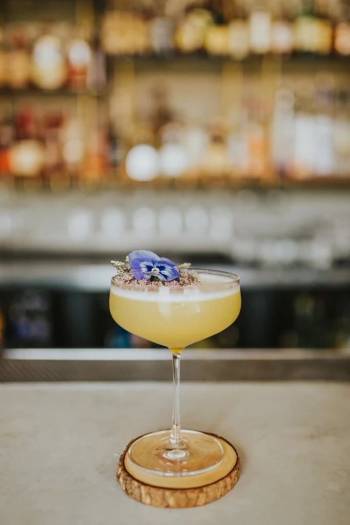 Garnished pale yellow cocktail in coupe glass on a wood coaster.