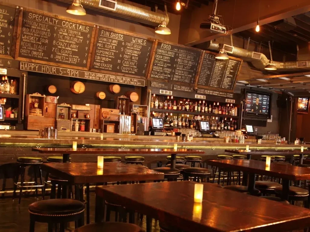 Dimly lit rustic-industrial bar with a stocked counter and menu chalkboards.