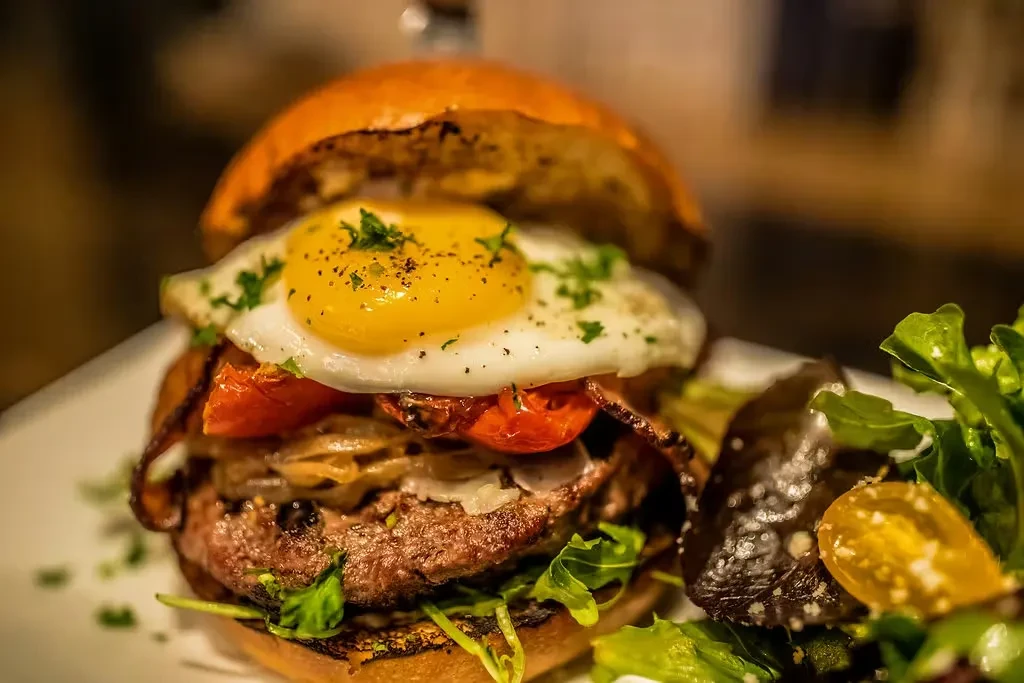 Close-up gourmet burger with fried egg, bacon, caramelized onions, side salad.