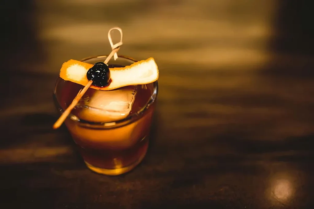 Close-up of an amber cocktail with ice, cherry, and orange peel.