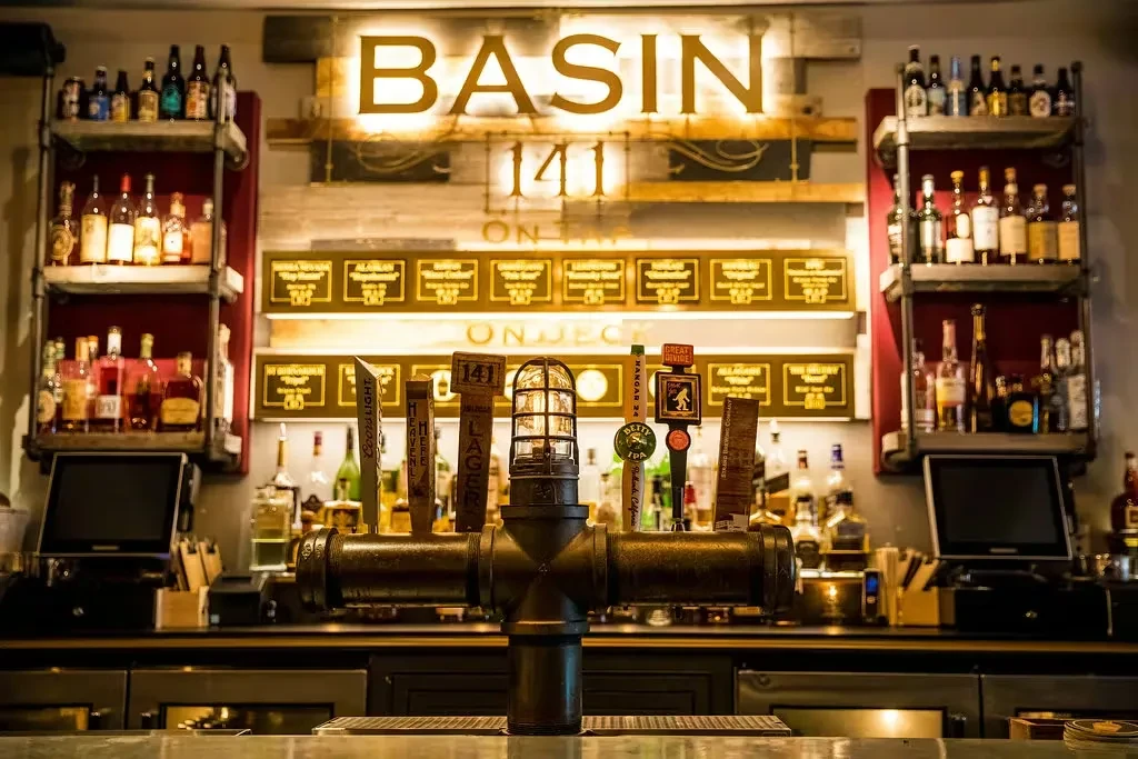 Industrial beer taps, bottles, and illuminated 'Basin 141' sign at a bar.
