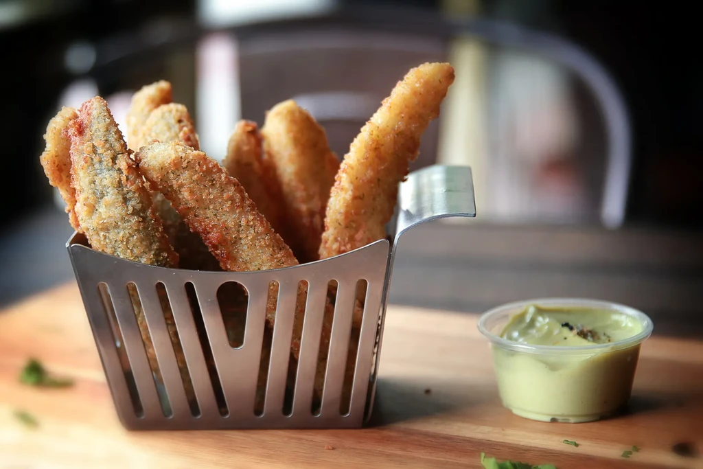 Golden-brown fried breaded sticks in a steel basket with green dipping sauce.