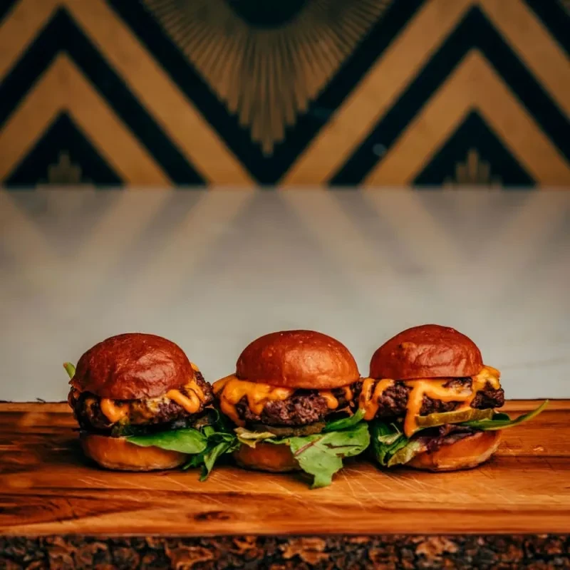 Three gourmet sliders with cheese, lettuce, and pickle on wooden board.