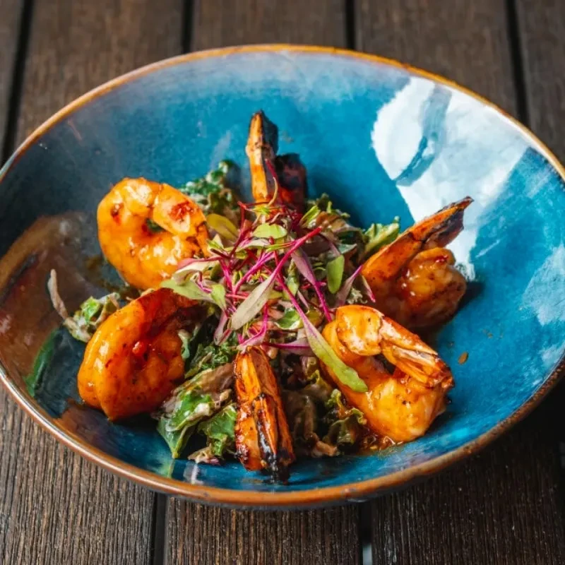 Five glazed shrimp with greens and microgreens in a blue bowl.