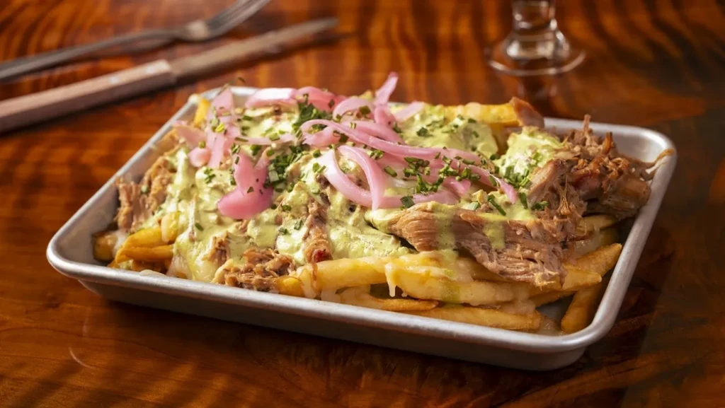 Loaded fries with shredded meat, green sauce, pickled onions, in silver tray.