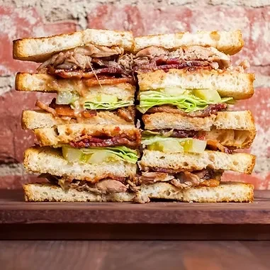 Toasted white bread sandwich with pulled meat, bacon, lettuce, pickles.