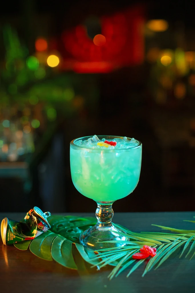 Light blue-green cocktail, crushed ice, candies, tropical leaves, crumpled can.