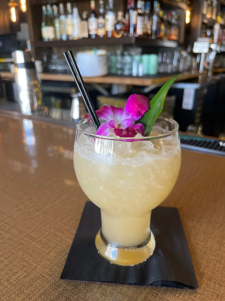 Frosty cocktail with magenta orchid, green leaf, and straws on bar.