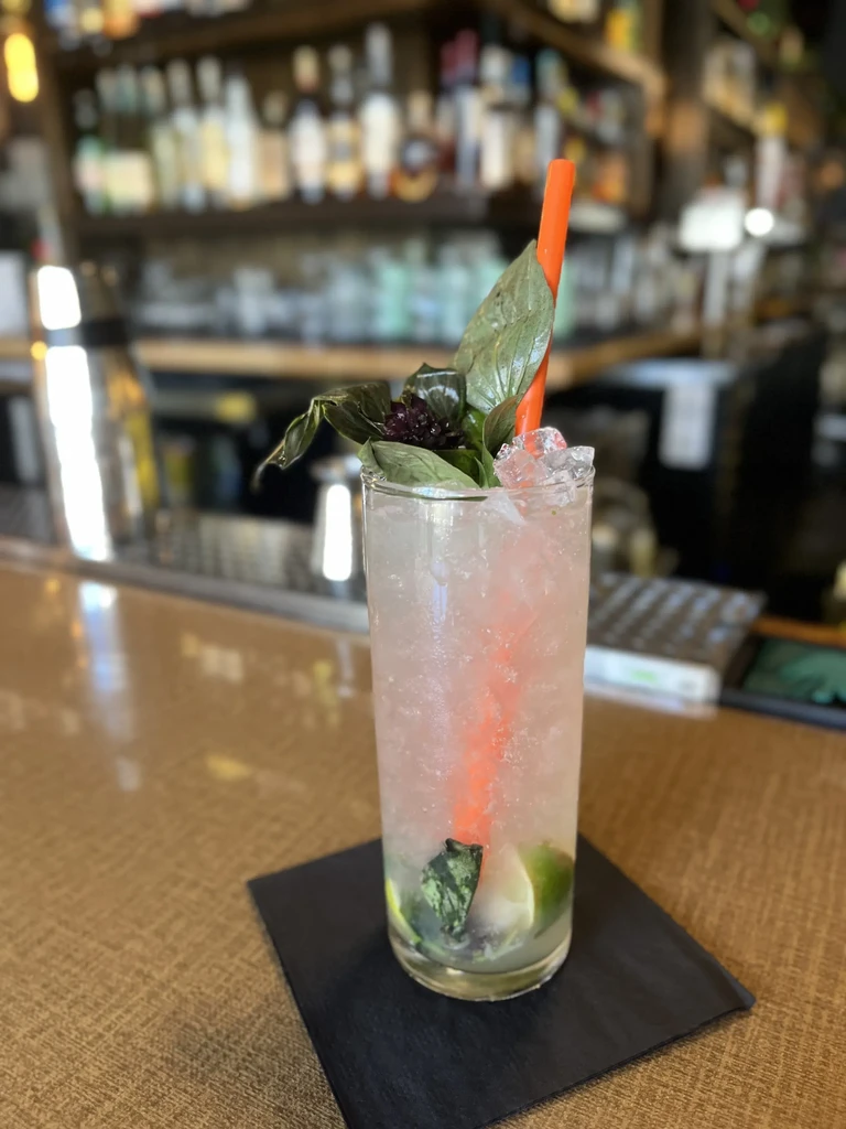 Cocktail with crushed ice, lime, basil, and orange straw on bar.