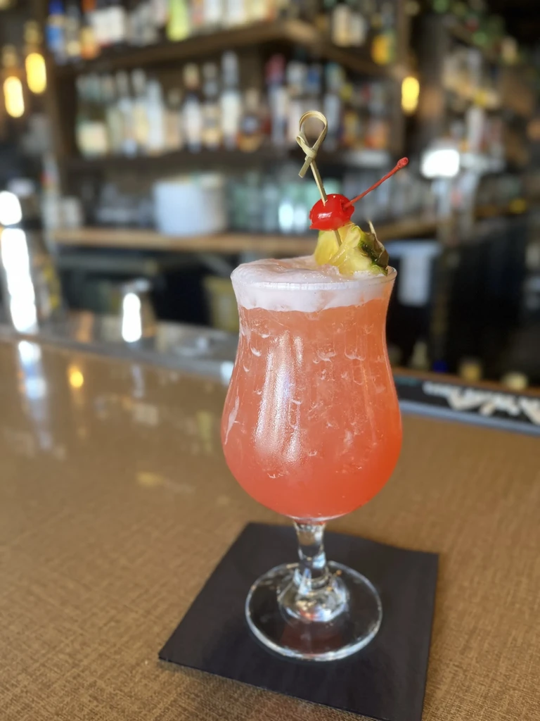 Reddish-orange cocktail in hurricane glass, garnished with pineapple and cherry.