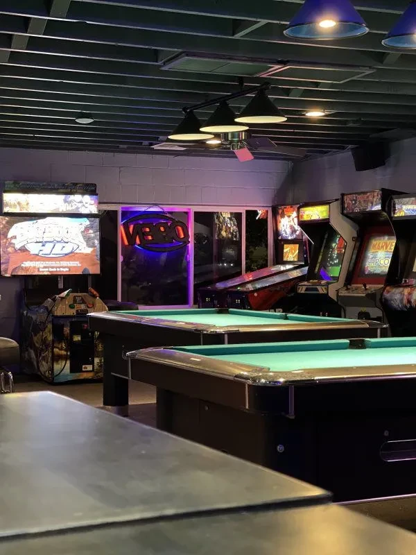 Two green pool tables and arcade machines in a dim game room.
