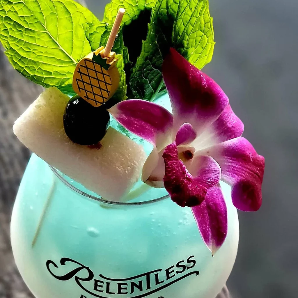Turquoise cocktail in 'RELENTLESS' glass, with fruit, mint, orchid garnishes.