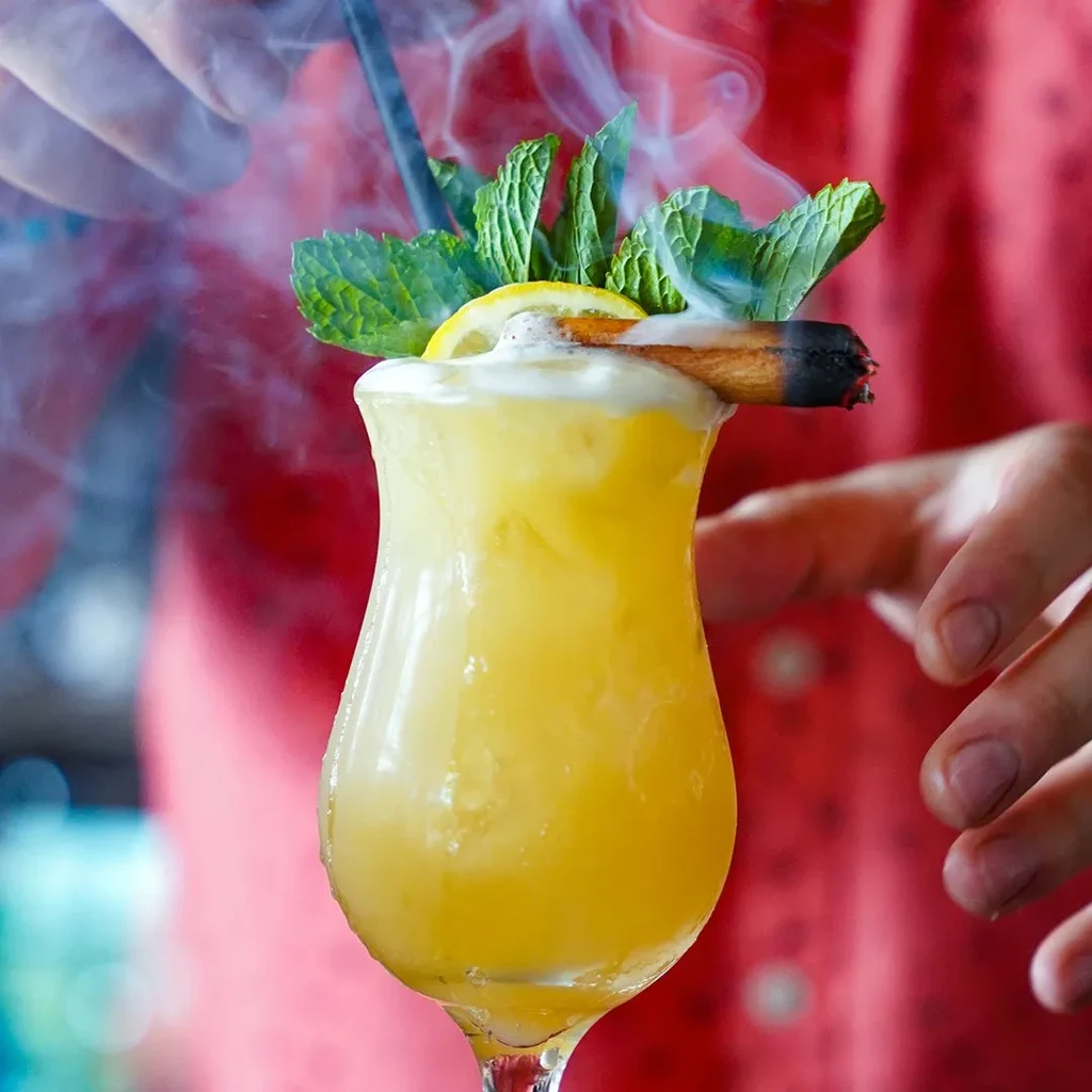 Yellow-orange cocktail in hurricane glass with foam, mint, and smoking cinnamon.