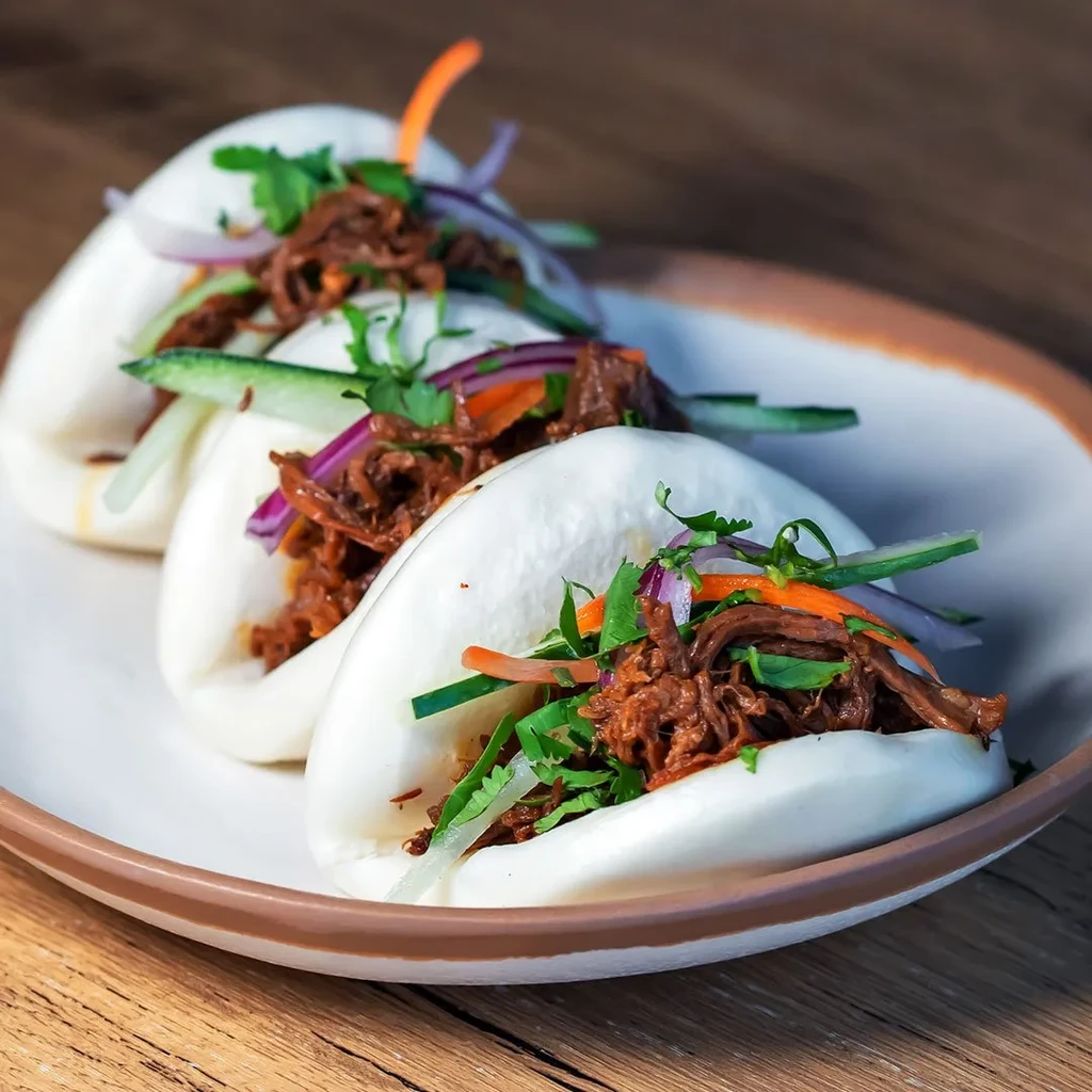 Three bao buns with shredded meat, red onions, cucumber, carrots, cilantro.