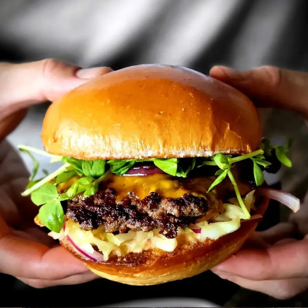 Two hands hold a burger with beef, cheese, sprouts, and red onion.