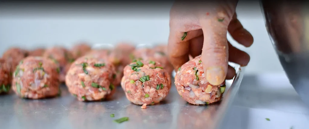 Hand shaping raw meatballs with herbs and vegetables on a steel surface.