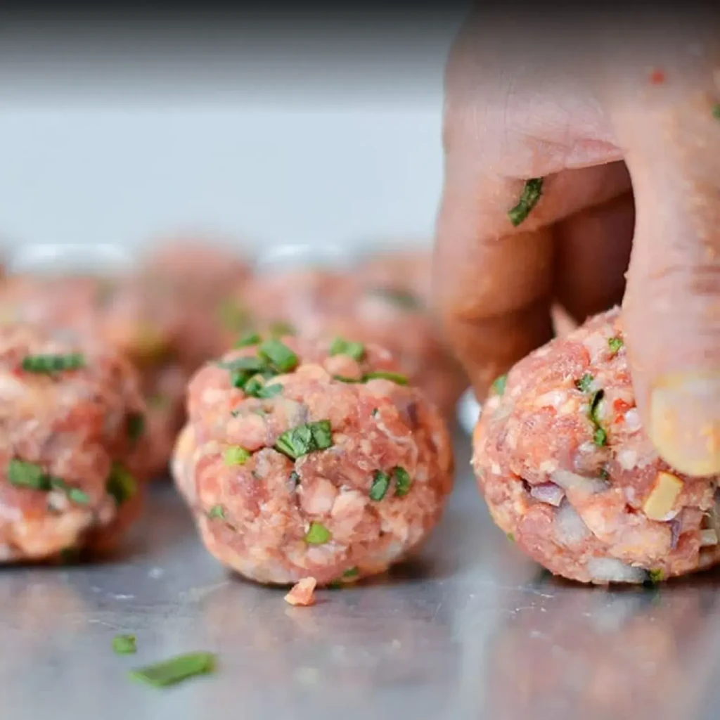 Hand preparing raw meatballs with visible red onions and green herbs.