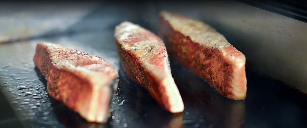 Three sizzling cuts of seasoned meat cooking on a dark griddle.