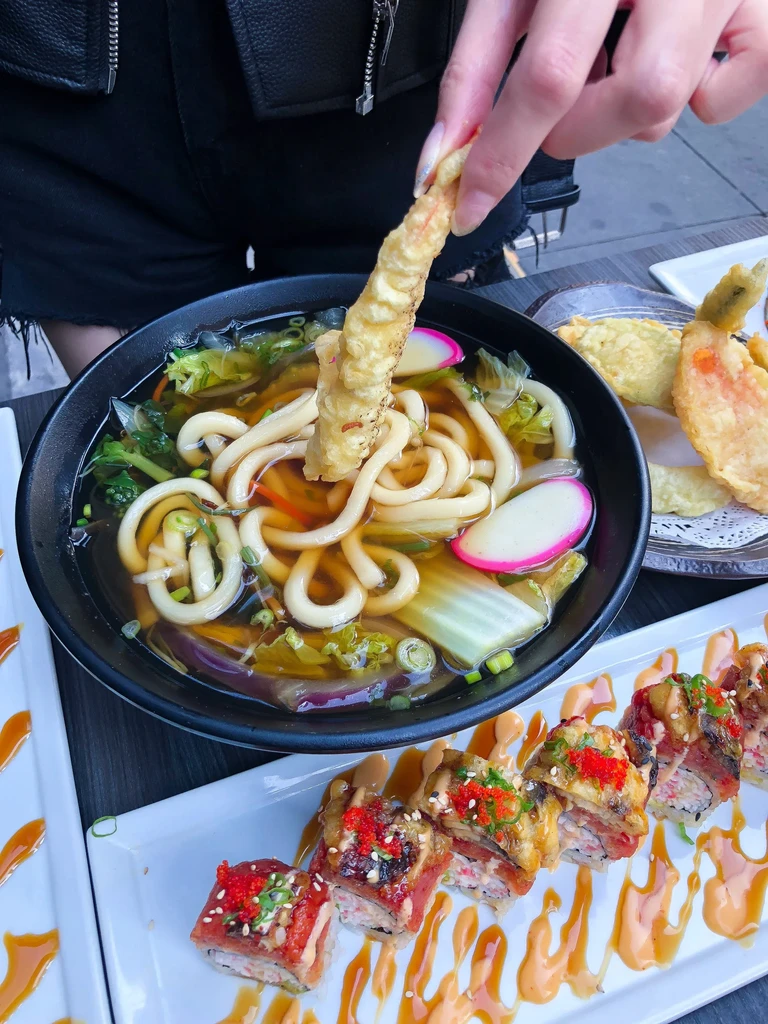 Close-up of hand dipping tempura into udon, beside garnished sushi.