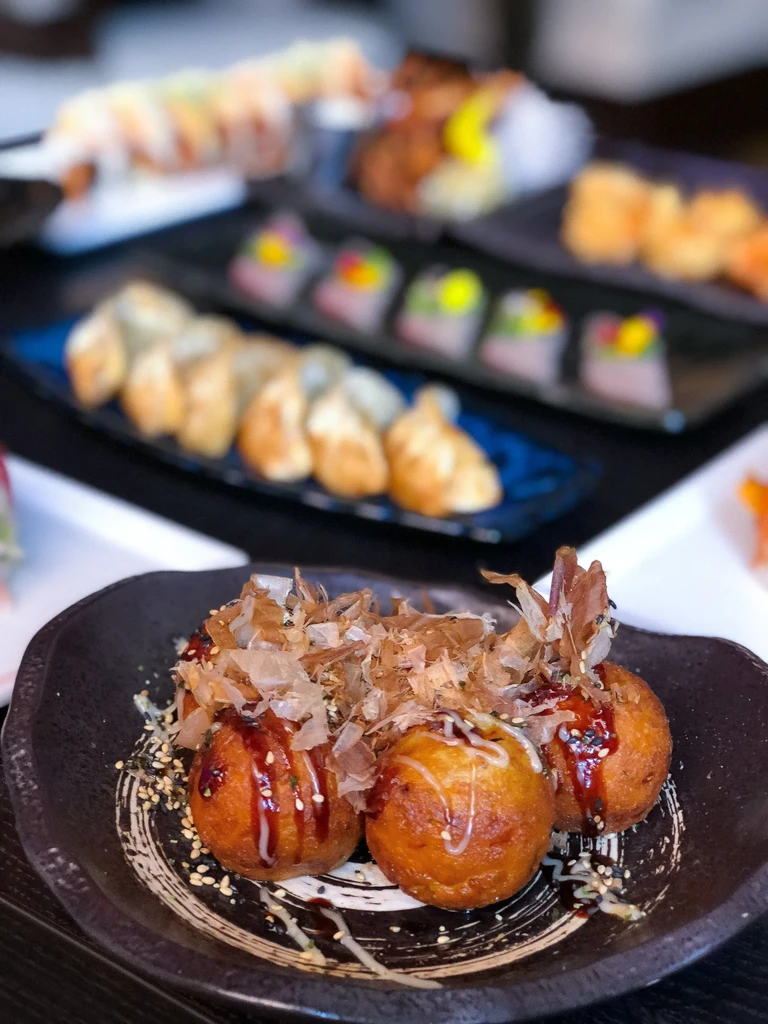 Plate of Takoyaki with sauce and bonito flakes, other Japanese dishes blurred.