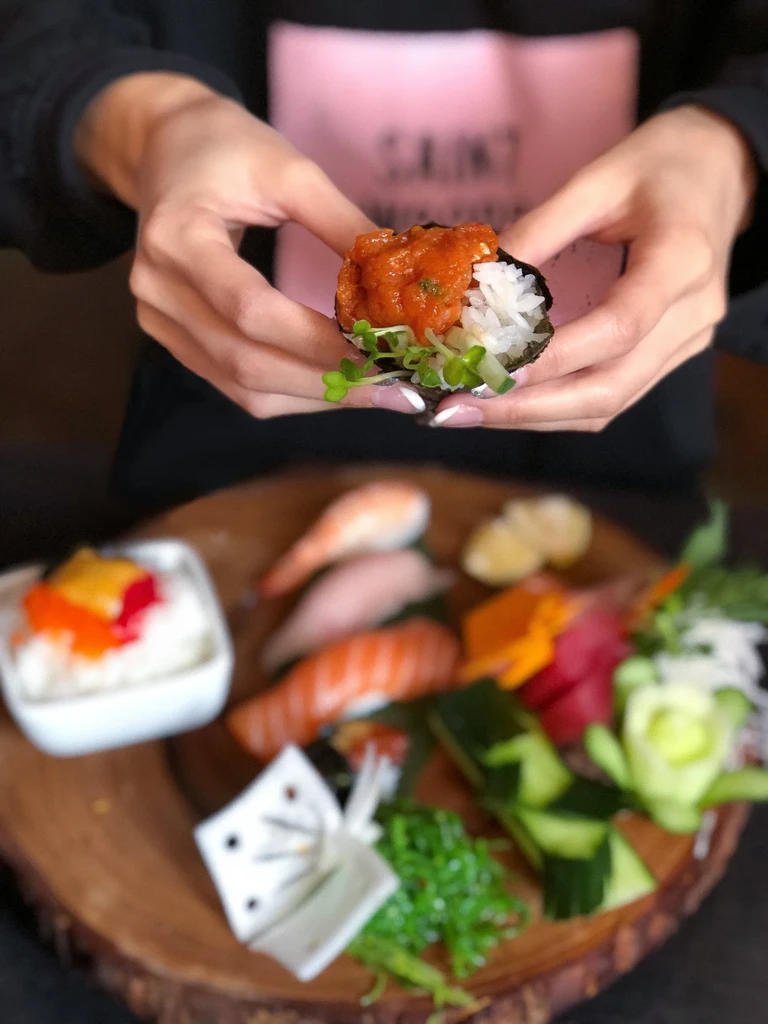 Hands holding a sushi hand roll; blurred sushi and sashimi platter behind.