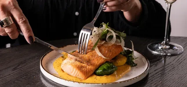 Close-up of person cutting cooked salmon, vegetables, and sauce.