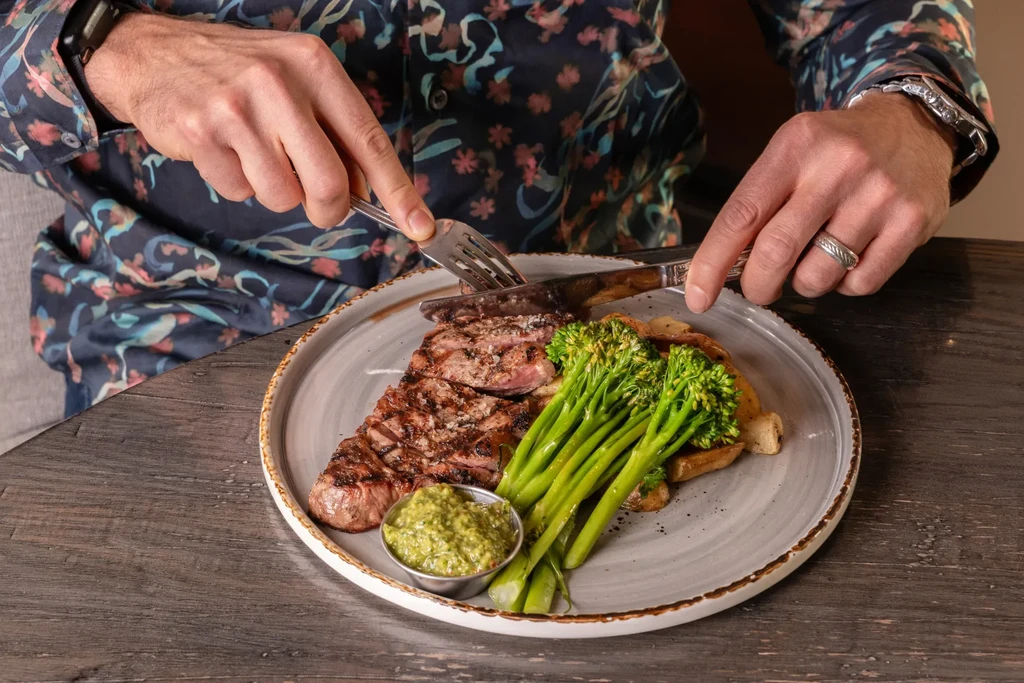 A person's hands cutting a grilled steak with broccolini and sauce.