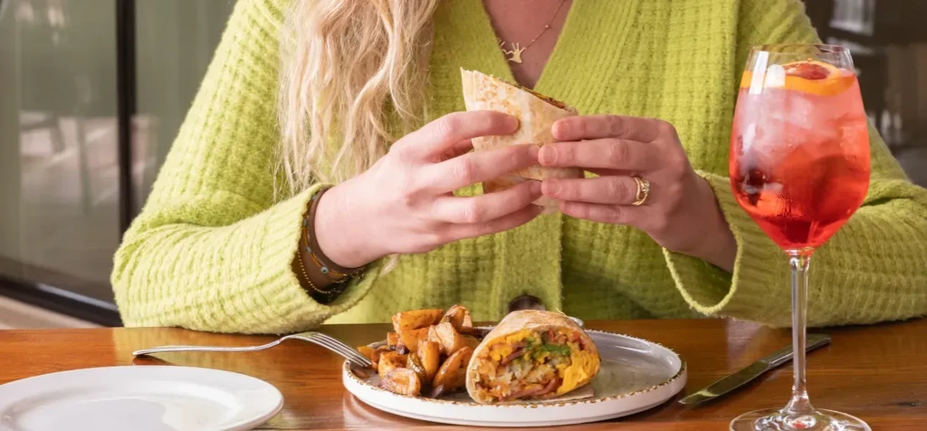 Hands holding a large wrap with roasted potatoes and a red cocktail.