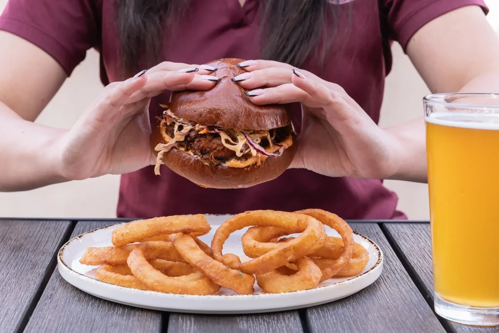 Large pulled pork burger held by hands, with onion rings and beer.