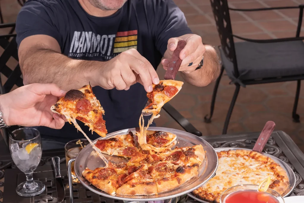 Close-up: Hands serving pepperoni pizza with cheese pull on outdoor patio.