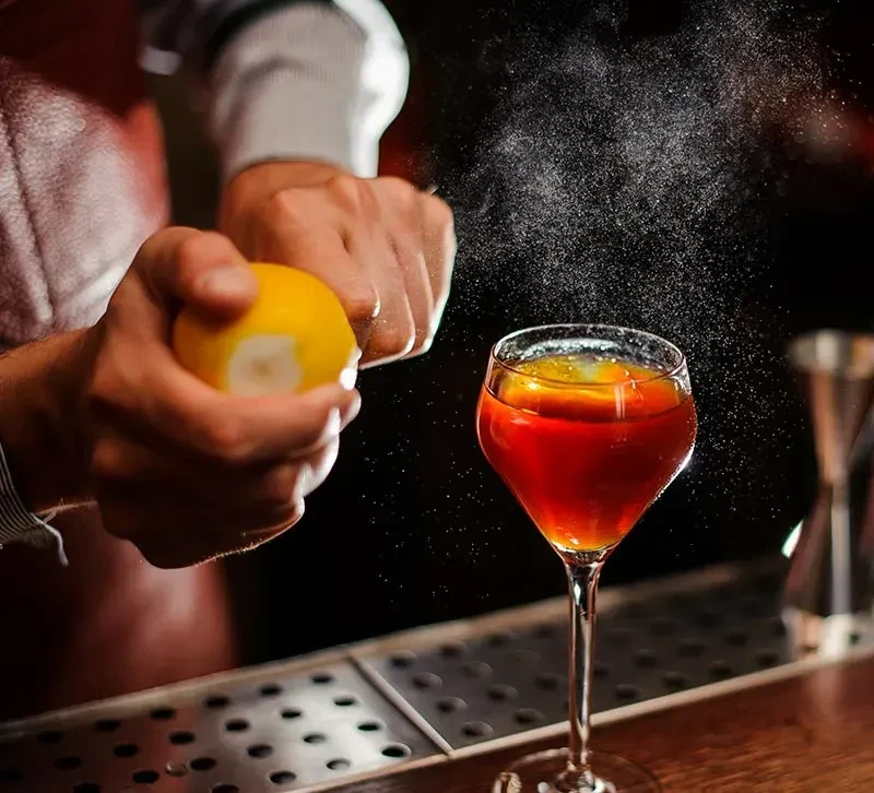 Bartender's hands squeezing lemon, expressing oils into a red cocktail.
