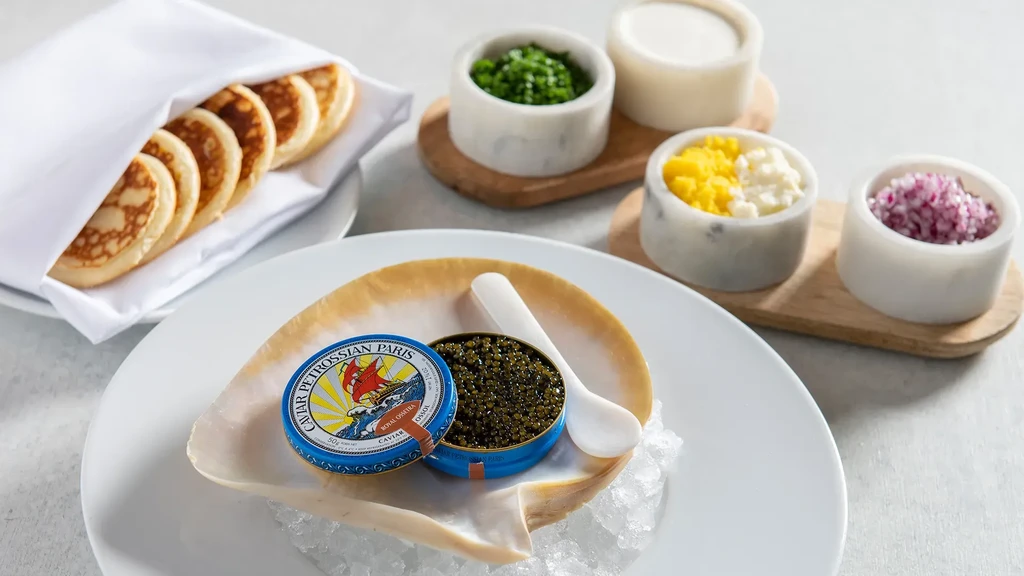 Open Petrossian Royal Ossetra caviar tin in seashell, blinis, garnishes.