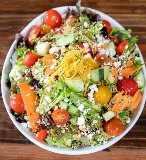 Overhead colorful salad with greens, tomatoes, cheese, seeds in white bowl.