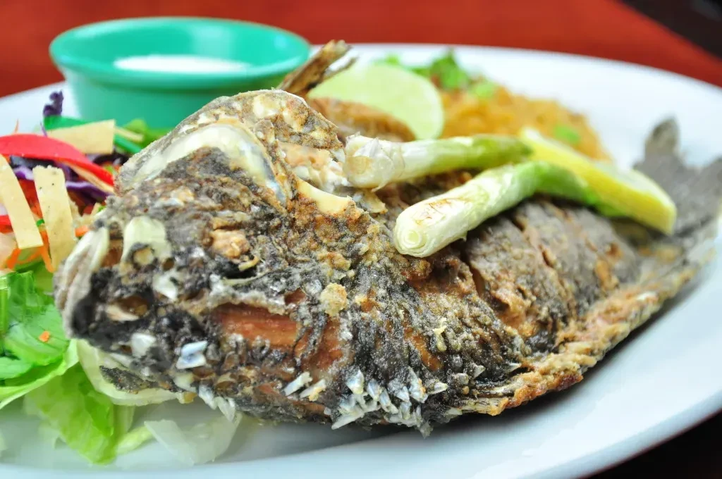 Crispy golden-brown whole fried fish with salad, rice, lime on white plate.