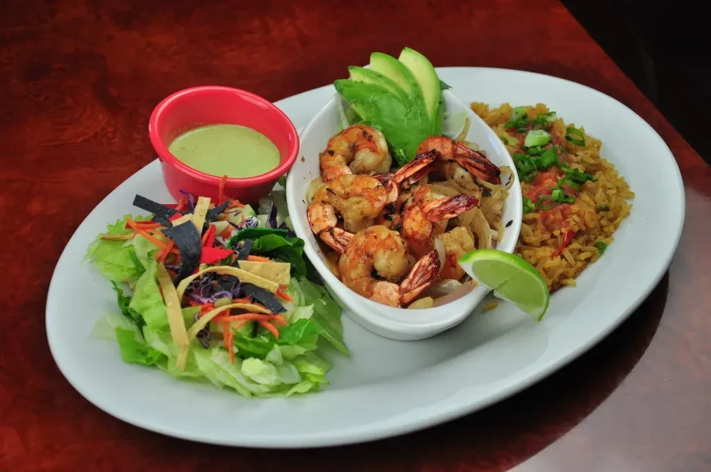 Grilled shrimp, salad, rice, and avocado crema on a red table.