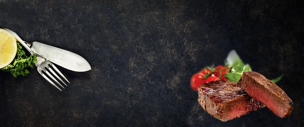 Medium-rare steak, cherry tomatoes, basil, lemon, and cutlery on dark background.