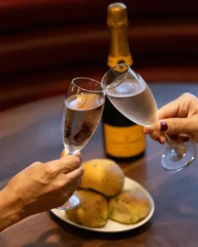 Two hands clink champagne flutes; bread rolls and bottle visible.