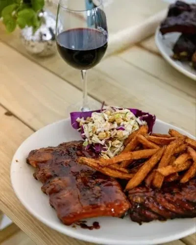BBQ ribs, sweet potato fries, coleslaw, and red wine on table.