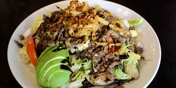 Top-down mixed green salad with meat, avocado, tomatoes, crispy onions.
