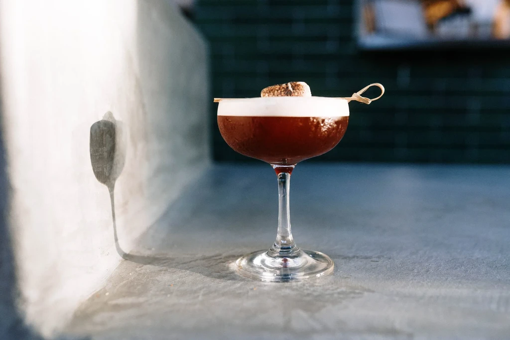 Dark cocktail with white foam and toasted marshmallow in coupe glass.