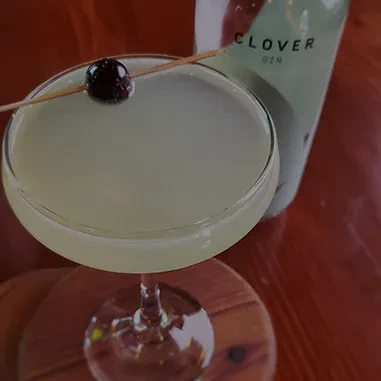 Pale green cocktail with cherry in coupe glass, partial Clover gin bottle.