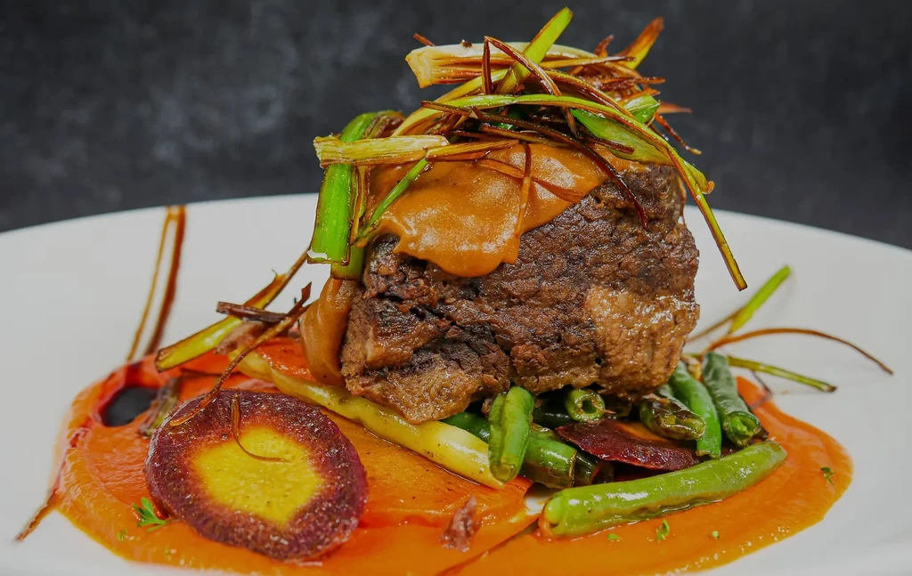 Meticulously plated braised meat, creamy sauce, colorful vegetables, orange purée.