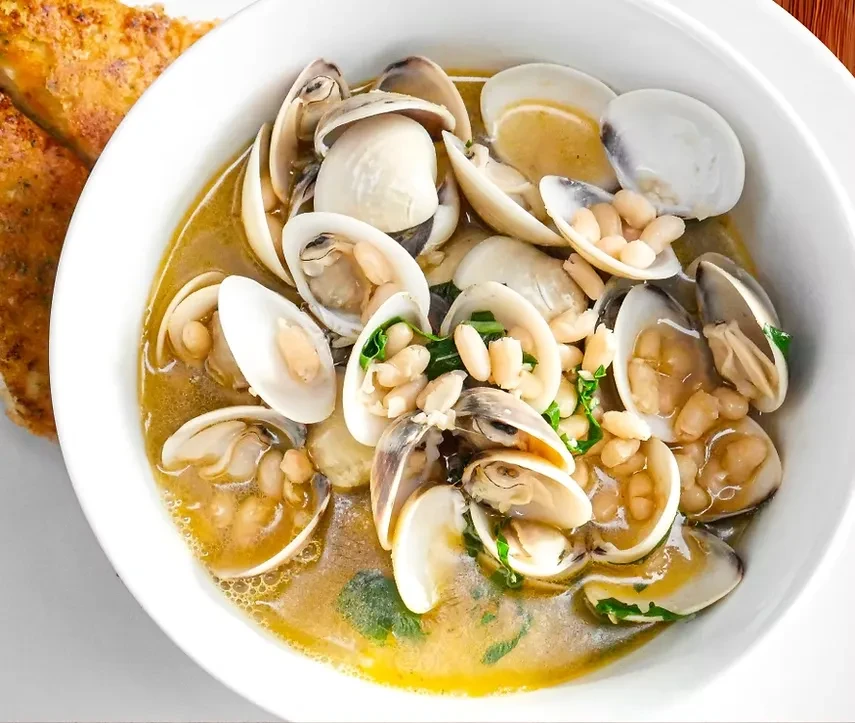 White bowl of clams, white beans, herbs in broth with toasted bread.