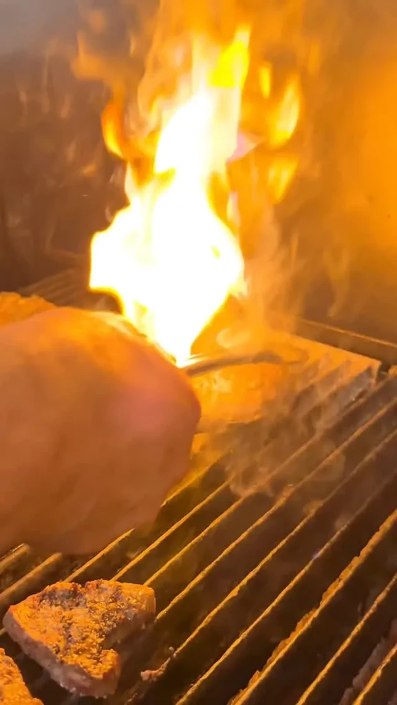 Hand uses tongs to grill meat over intense flames and smoke.