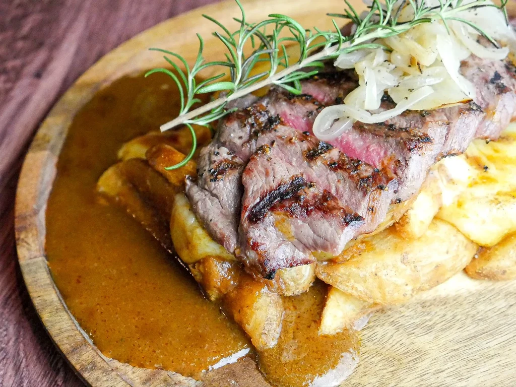 Sliced medium-rare steak with onions, rosemary, potatoes, and gravy on wood.